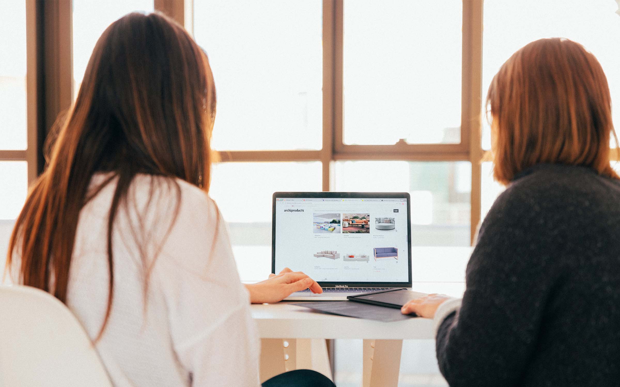 Two women sit with their backs to the camera collaborating on one shared computer