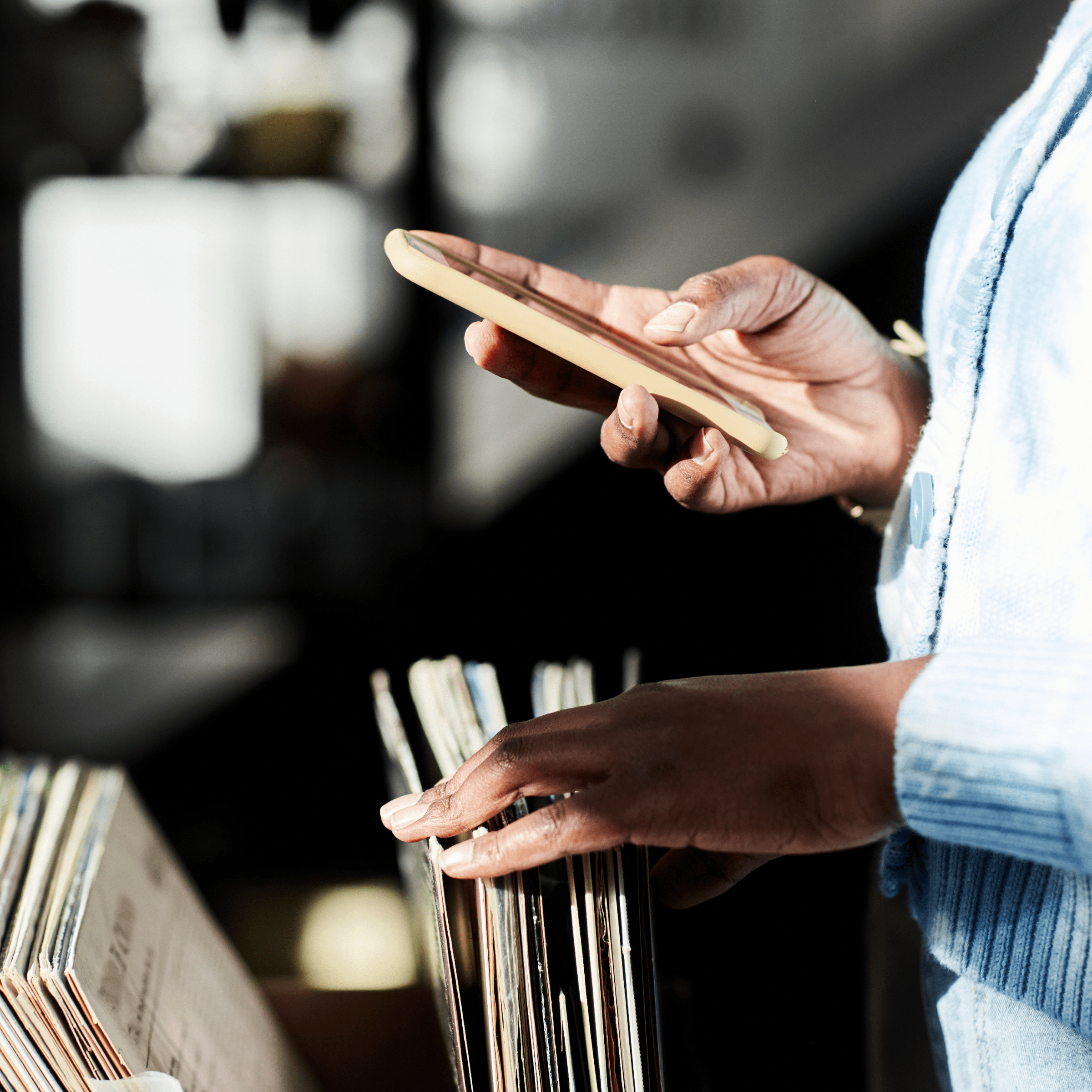 A close-up of a person browsing through a collection of vinyl records while holding a smartphone in their other hand. The person is wearing a light blue sweater, and the setting appears to be a record store or a similar environment with a nostalgic ambiance. The image captures the act of searching for music while possibly researching or cataloging information on the phone. The focus is on the hands, with the background softly blurred.