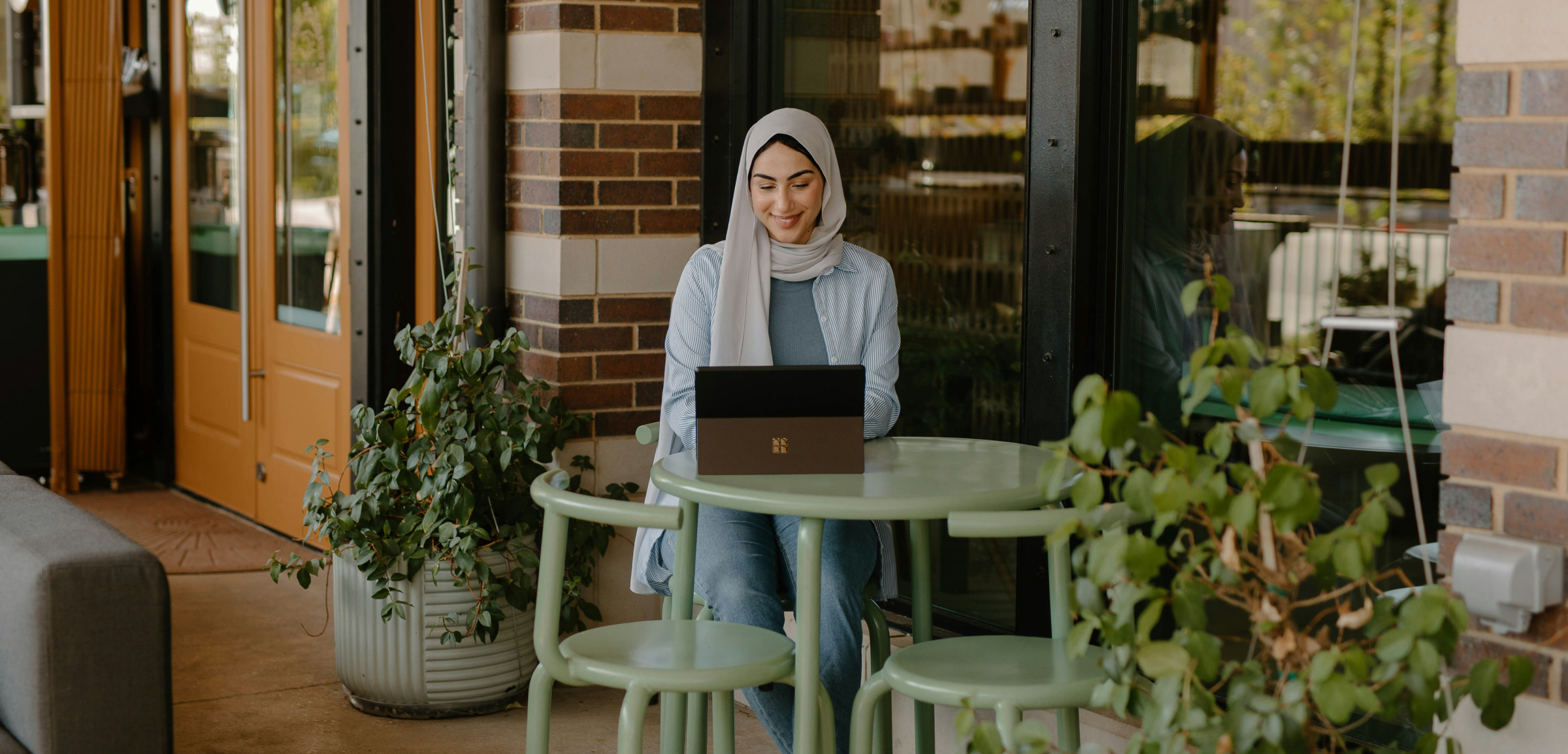 A woman wearing a hijab is sitting at an outdoor café, using a laptop. She is seated at a light green table with matching chairs, surrounded by potted plants. The setting is relaxed and pleasant, with warm natural light, a brick wall, and large windows in the background. The woman is smiling as she works, suggesting a comfortable and productive environment, blending nature with a modern workspace.