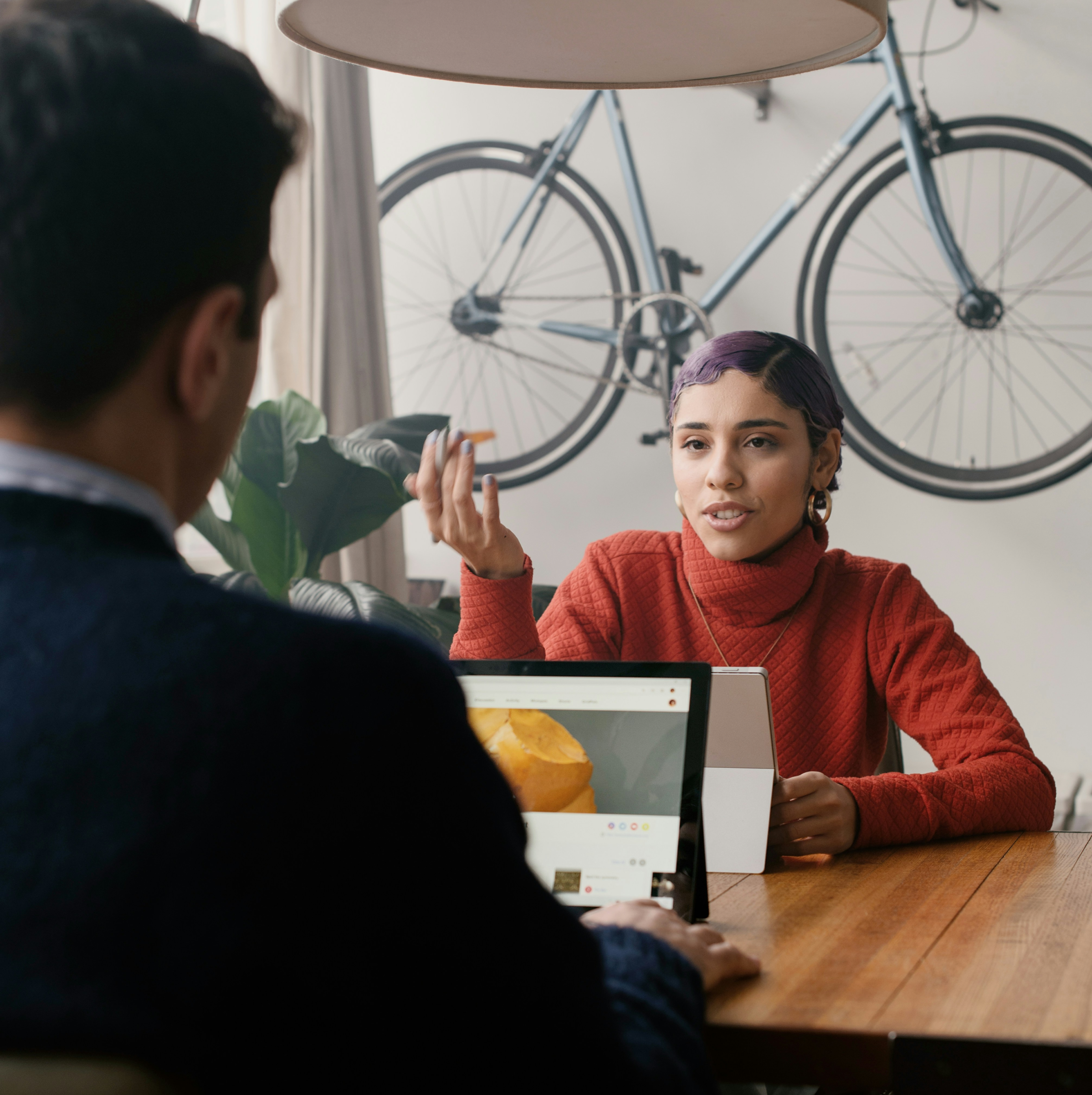 Two people are engaged in a discussion at a wooden table in a modern indoor setting. The person facing the camera is a young woman with short, purple hair, wearing a red sweater, gesturing as she speaks. The other person, with their back to the camera, is using a laptop that displays a website. A bicycle is mounted on the wall behind them, adding a creative and contemporary touch to the space. The environment is casual and relaxed, suggesting a collaborative conversation or brainstorming session.