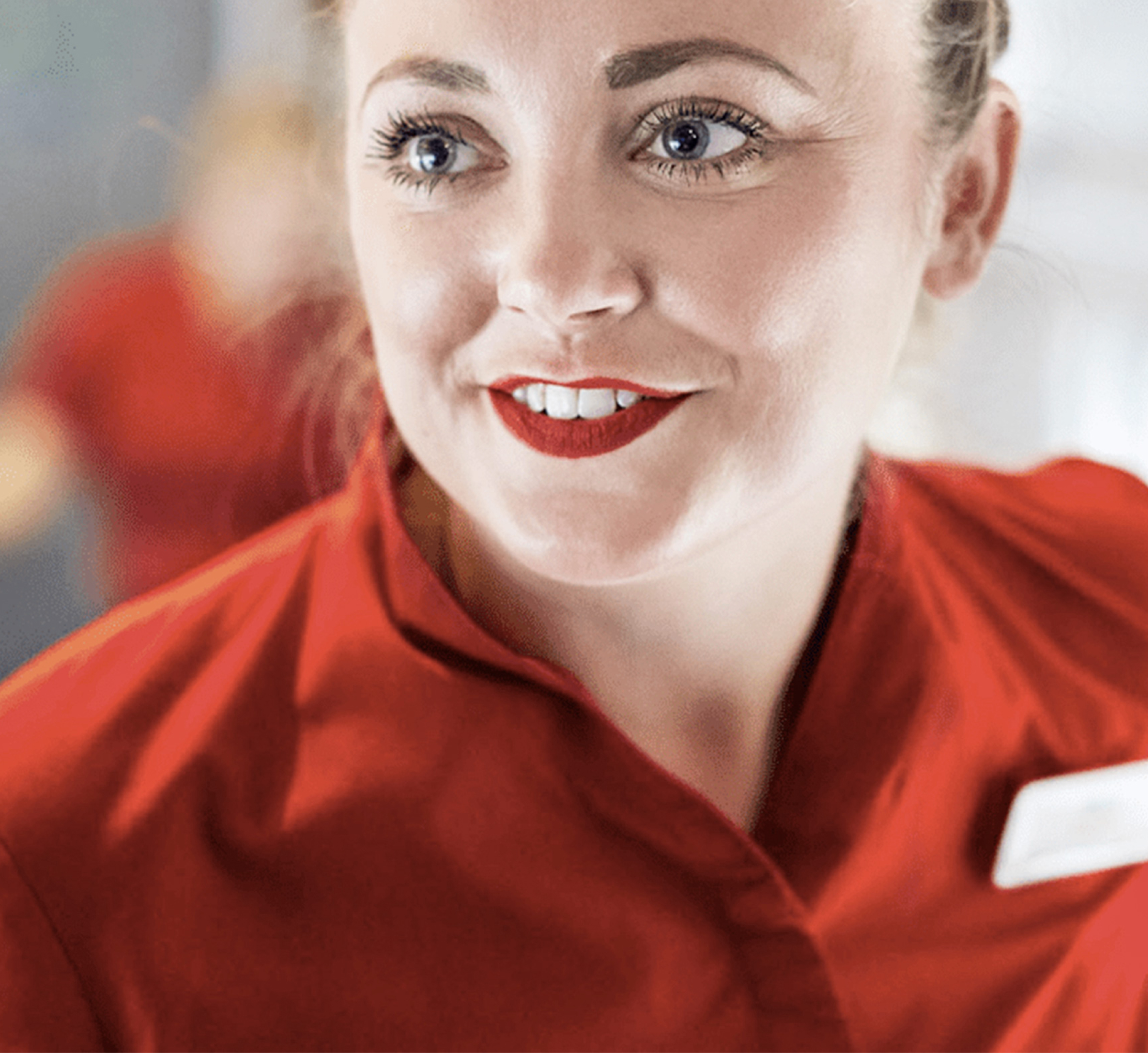 A close-up of a smiling female employee wearing a red uniform, with a friendly and welcoming expression. The background is softly blurred, focusing attention on her face.