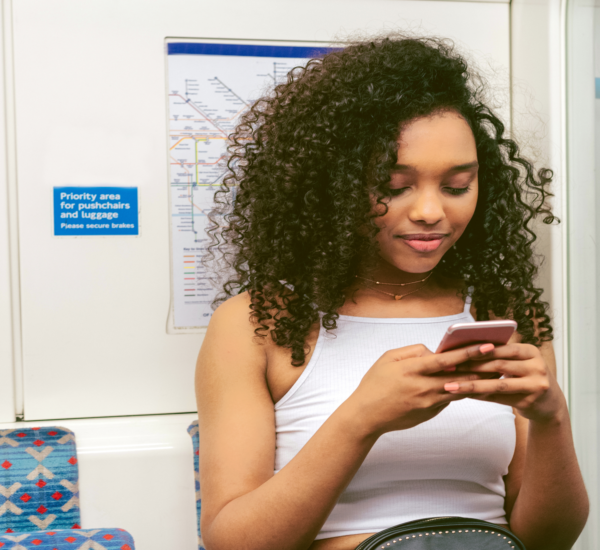 "A young woman with curly hair sitting on public transportation, looking at her phone. Behind her, a map of the subway system is visible, along with a sign designating the area for pushchairs and luggage. She appears relaxed, enjoying her ride.